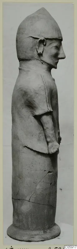 Terracotta statue of a standing figure in side view, with a tall pointed head covering and arms held against the body