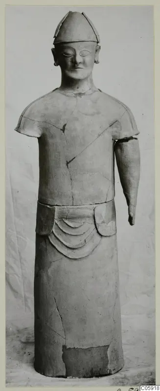 Terracotta standing figure with a tall conical cap and one arm missing, photographed against a draped cloth