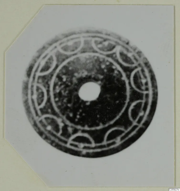 Flat disc-shaped object with a central hole, decorated with concentric rings and a ring of semicircular arcs, shown on a card mount.