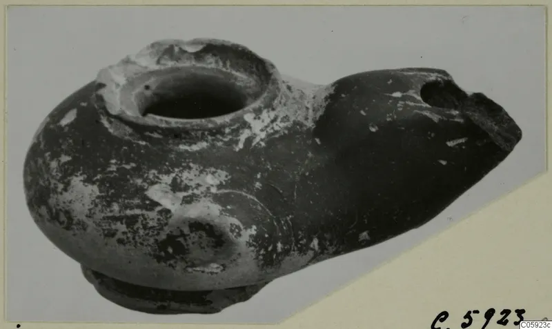 Terracotta oil lamp from Idalion with a broken nozzle, photographed at an angle against a grey backdrop