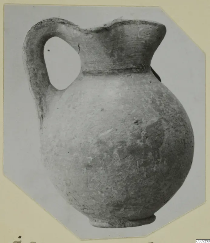 Round-bodied jug with a strap handle and flared mouth, shown on a pale card mount with cut corners