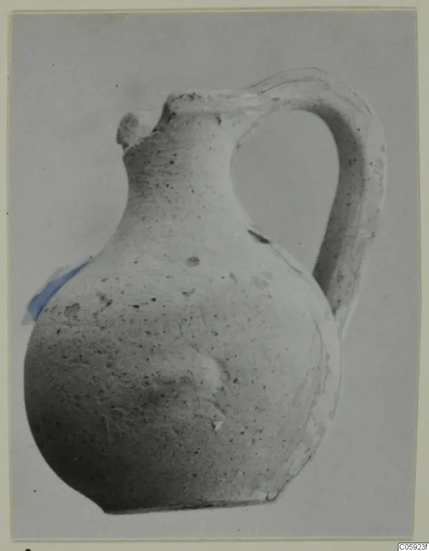 Single-handled pottery jug with a globular body, narrow neck and broken lip, pale surface with dark speckles.