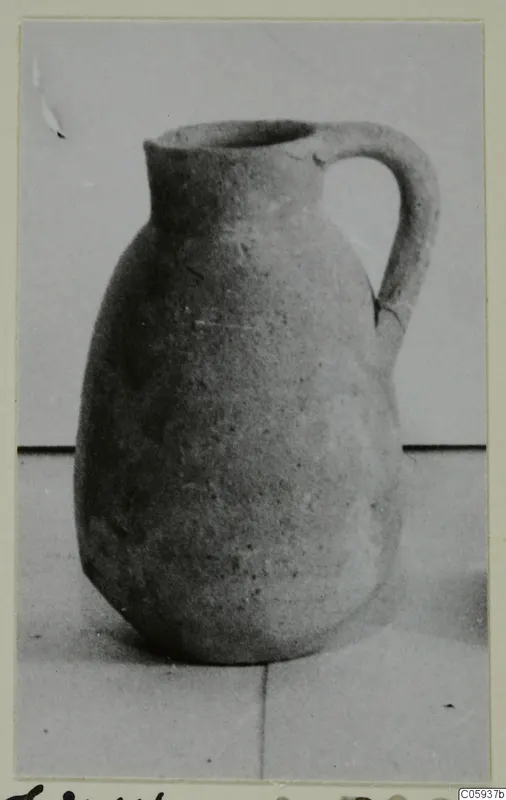 Pottery jug with a single loop handle and a wide mouth, shown on a pale surface against a grey backdrop.
