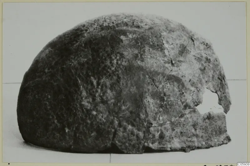 Corroded metal object with a domed shape and a broken opening on one side, photographed against a pale background