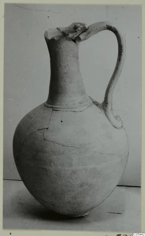 Pottery jug with a tall neck, single loop handle and broken rim, photographed against a grey backdrop.
