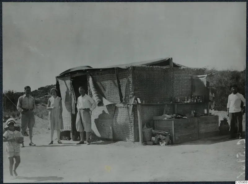 People standing in front of a reed-walled tent at Oura, with a wooden counter beside it.