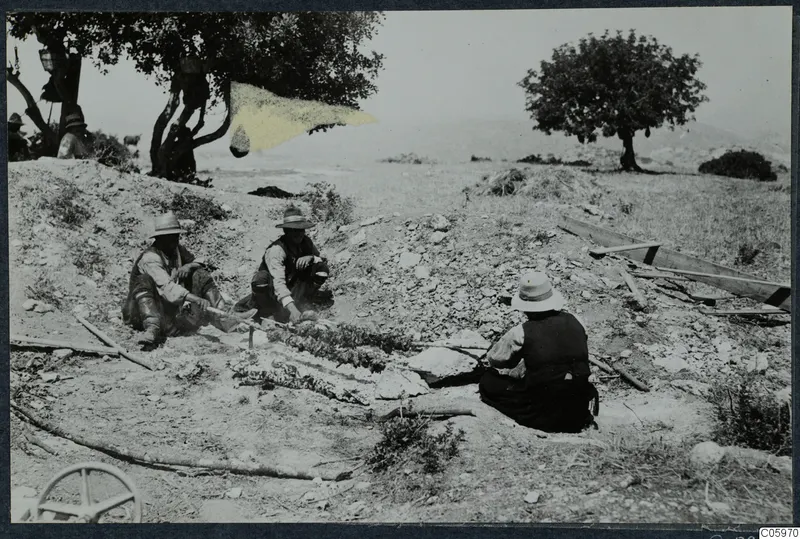 Men sit on a stony slope roasting food on skewers at Vouni, under a tree
