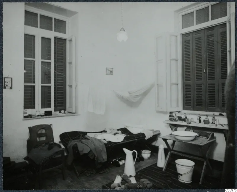 Interior of a room in Nicosia with a camp bed, a folding washstand and tall shuttered windows.