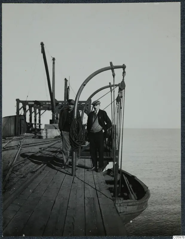 Two men standing on a wooden pier at Karavostasi, with a small boat and the sea beside them.