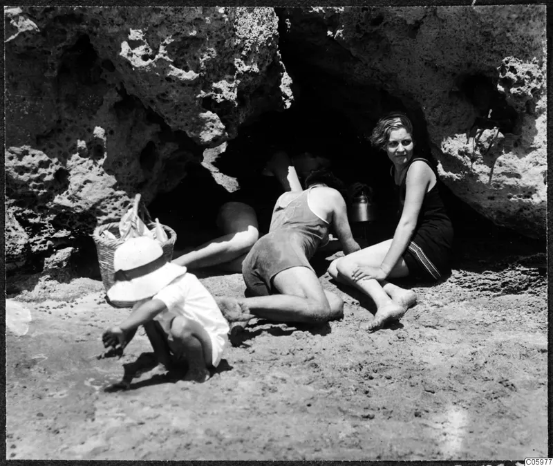 Four people at the mouth of a rock cave on a sandy beach, a woman in a dark swimsuit seated at the entrance and a child crouching in front