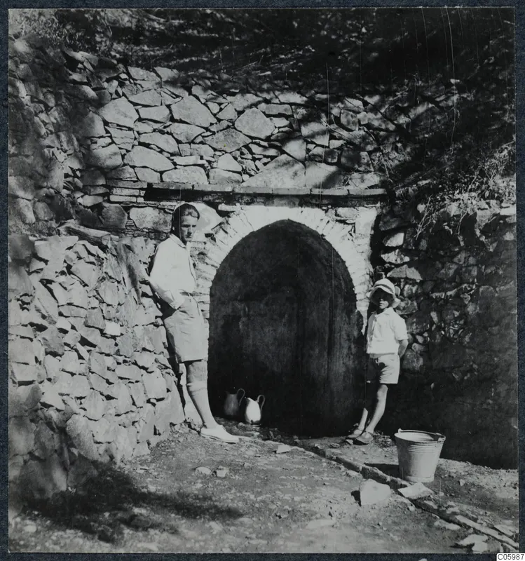 Man and boy standing beside a stone-arched water source at Machairas