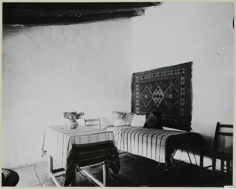 Interior of a whitewashed room in Machairas, with a striped-covered bench, a wall rug and a table holding a vase of flowers.