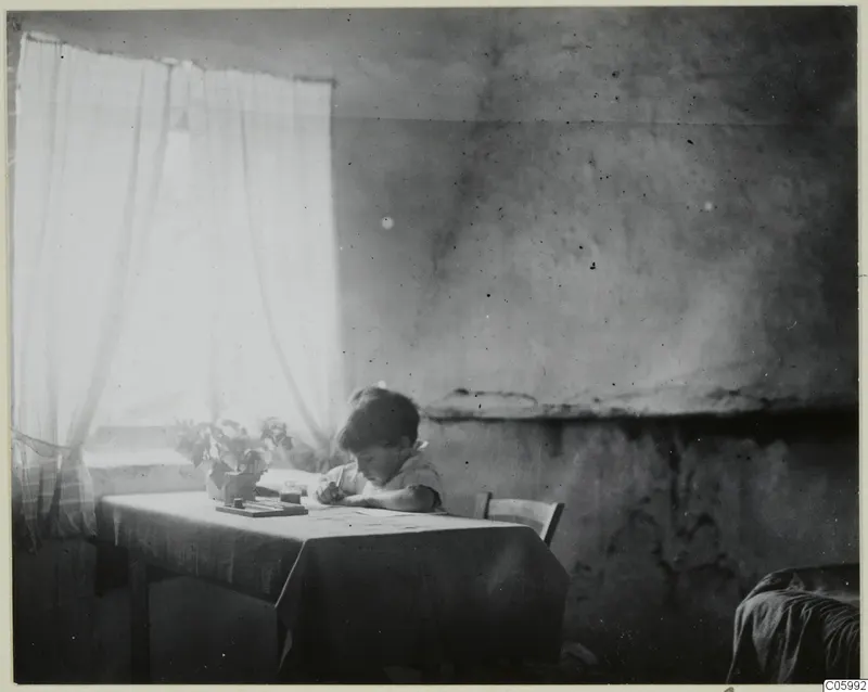 Interior at Machairas: a small boy writes at a table beside a window with sheer curtains.