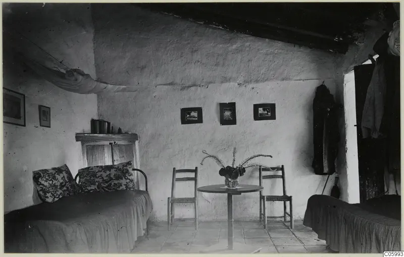 Interior of a whitewashed room at Machairas with two metal beds, a round table, two chairs and framed pictures on the wall.