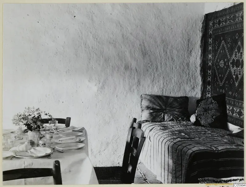 Interior at Machairas with a bed under a hanging carpet and a formally laid table with a white cloth.