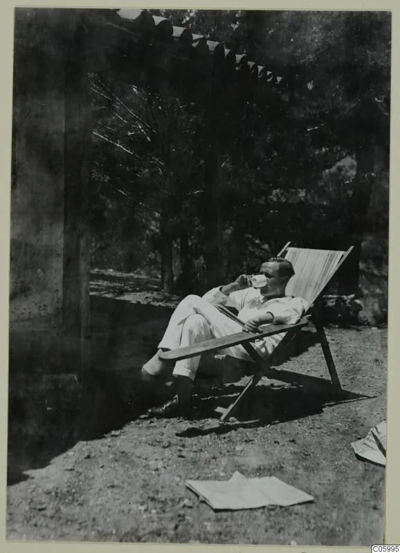 A man in a white suit reclines in a wooden deck chair under trees at Machairas, drinking from a cup.