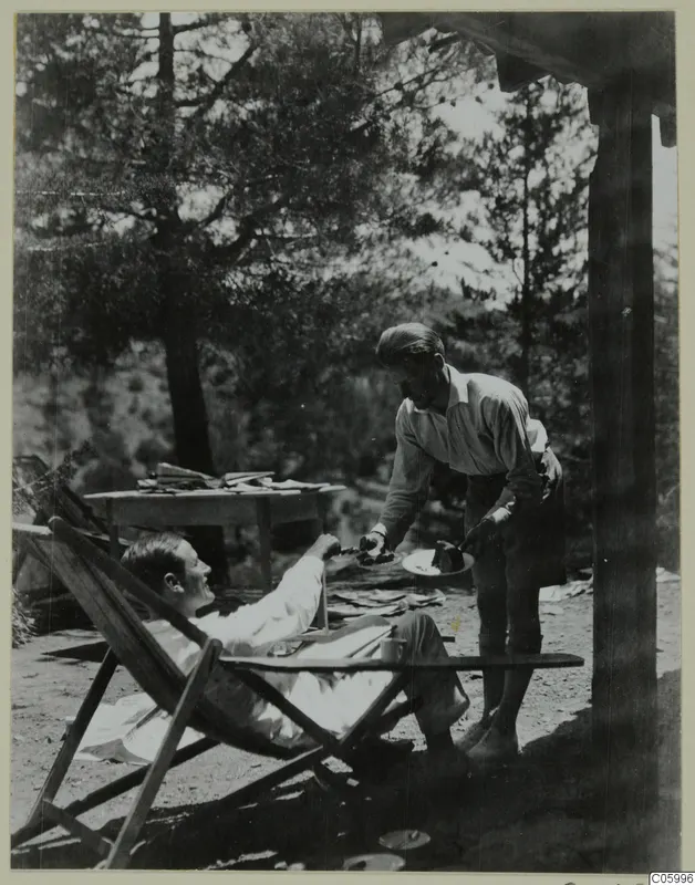 Two men on a terrace at Machairas: one reclines in a deck chair while the other offers him a slice of cake.