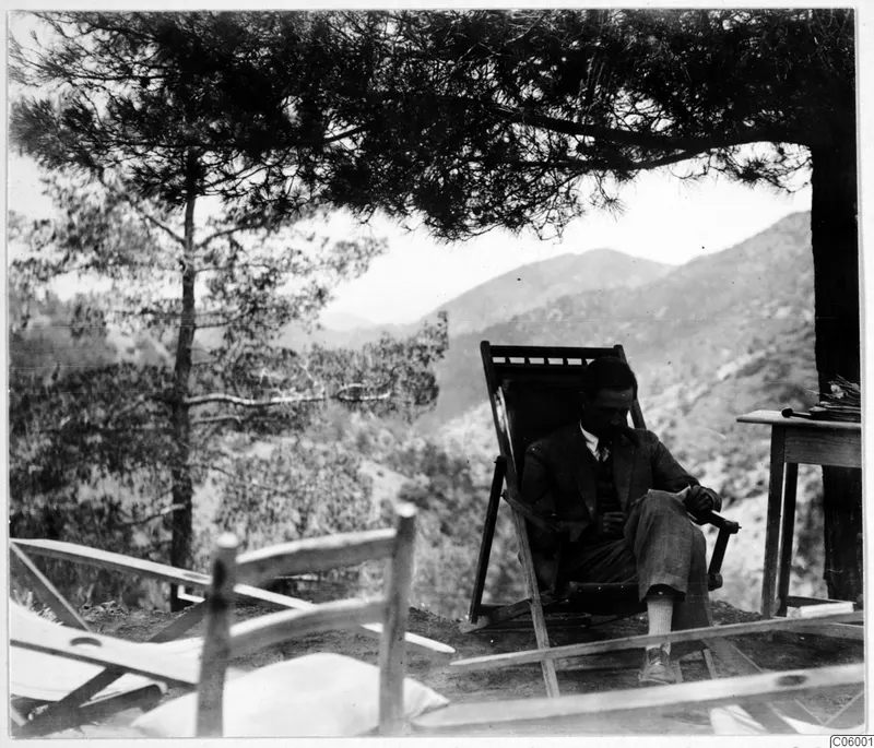 A man, probably Einar Gjerstad, rests in a wooden deck chair on the terrace at Machairas, with pine branches and hills behind him.