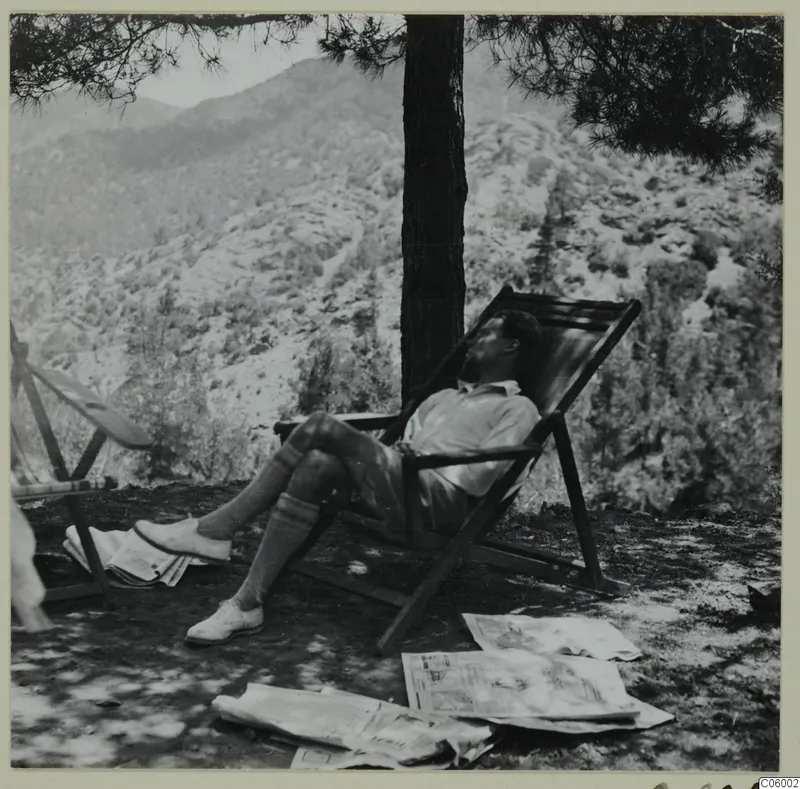 Alfred Westholm resting in a wooden deck chair under a pine tree at Machairas, with newspapers on the ground.