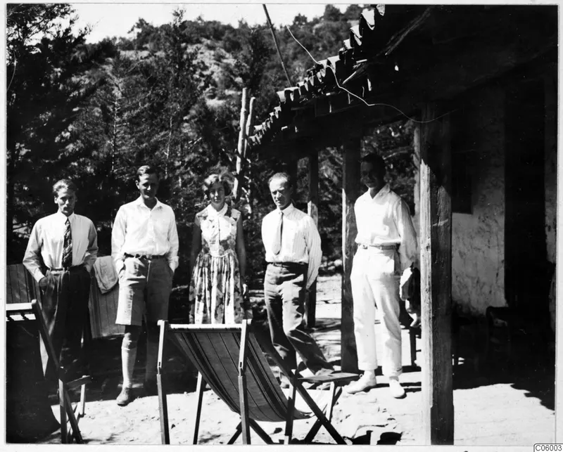 Five people stand together on a sandy terrace outside the expedition house at Machairas, with a wooden deckchair in front.