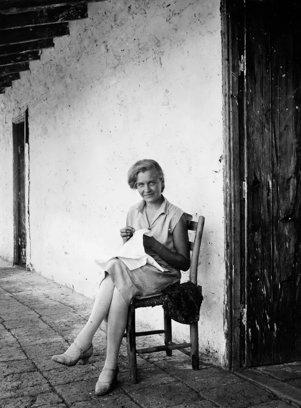 Woman sewing while seated on a wooden chair on a paved terrace beside a whitewashed wall and a wooden door.