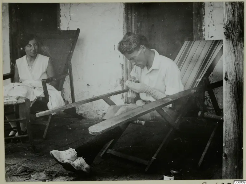 Man darning a sock on a stone terrace, with a woman seated in a deck chair beside him