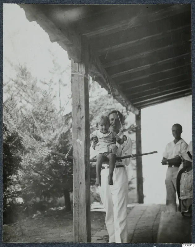 Man in white holds a small boy on a wooden swing bar under a timber terrace roof at Machairas; two men stand at right.