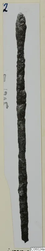 Long, thin, corroded bronze rod with a rounded top, photographed vertically against a pale card mount