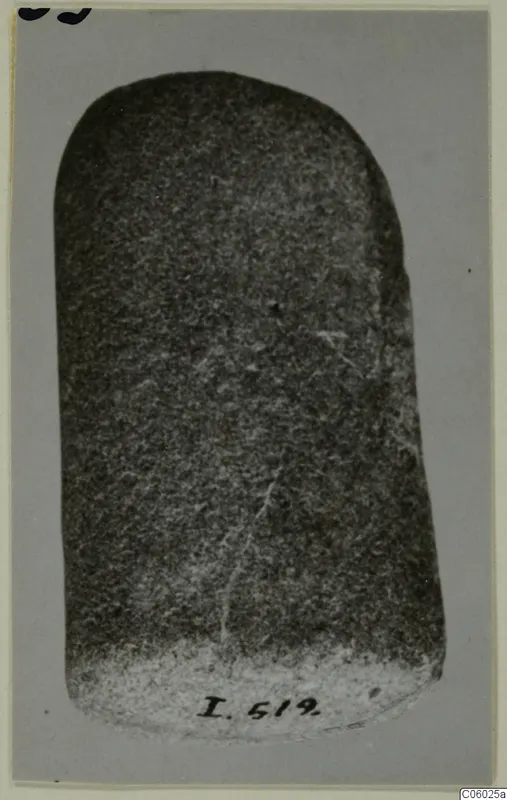 Pestle, a dark stone object with a rounded top, photographed upright against a grey backdrop