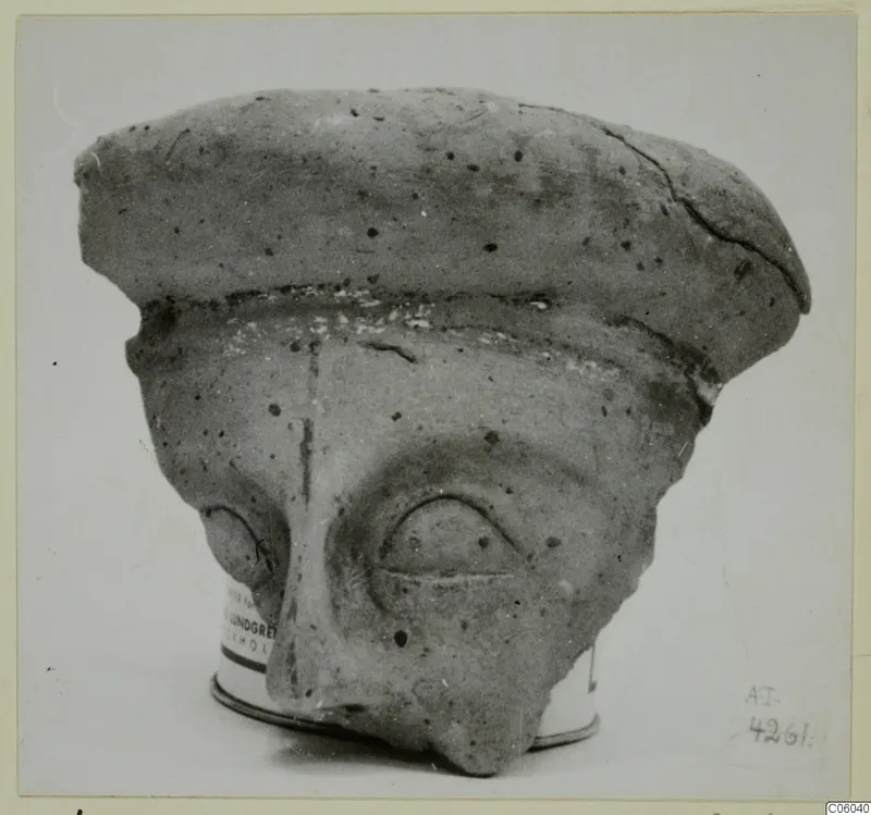 Terracotta face fragment with a flat, cap-like top, a large relief eye and a broken lower face, shown on a tin base.