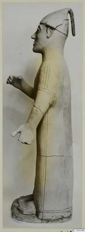 Terracotta standing figure in profile facing left, with a tall cap and incised lines on the robe and arm.