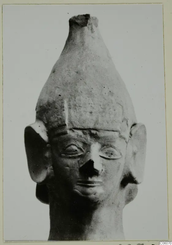 Terracotta head of a figurine with a tall conical headdress, large flat ears and a broken nose