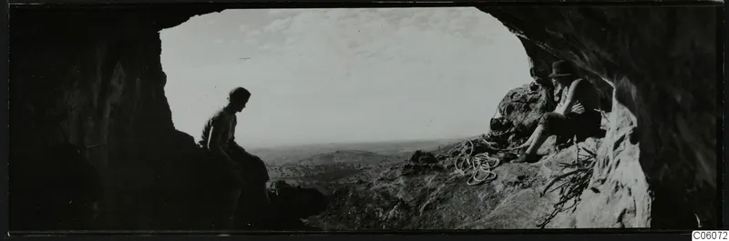 Two people seated at the mouth of a cave at Fylleri, Kythrea, with rolling hills in the distance.