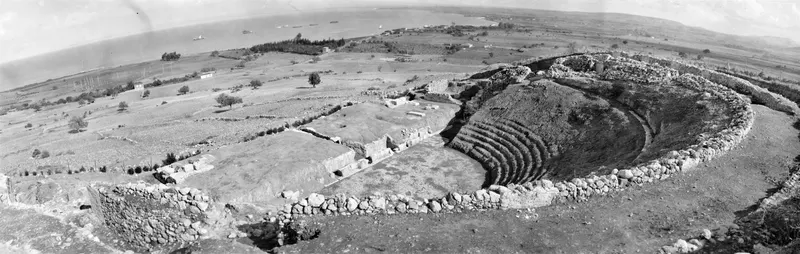 Panoramic view of the theatre at Soli, with curved stone seating rows, a stone wall and the sea beyond.