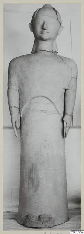 Standing terracotta figure with arms at its sides, shown frontally against a plain backdrop, with visible restoration joins.