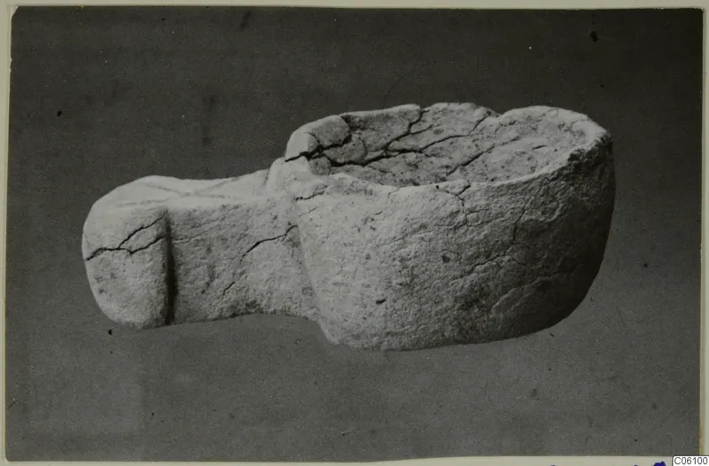 Stone bowl with a flat projection on one side, cracked, photographed against a dark backdrop