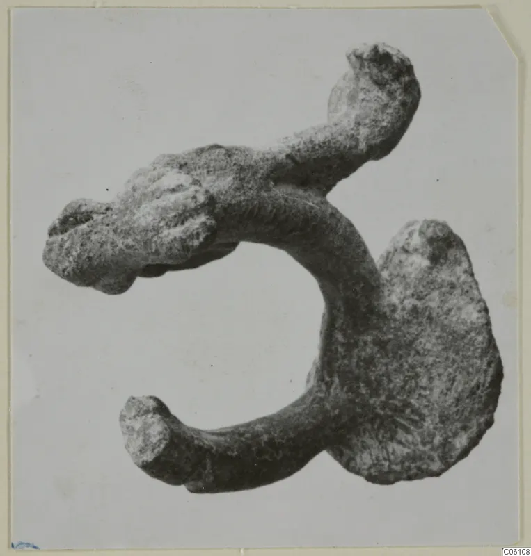 Corroded bronze fragment: a thick curved bar with knobbed ends and a broad flattened lobe.
