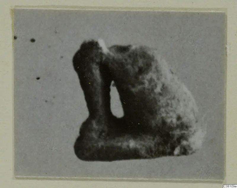 Blurred photograph of a small dark object with a vertical opening, from Ayia Irini, on a cream card mount