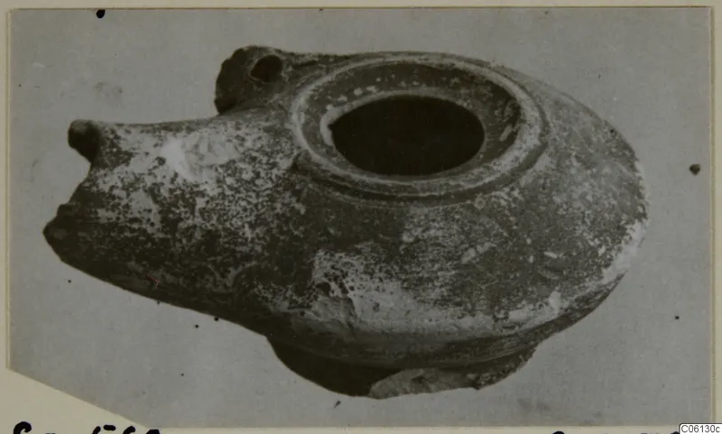 Terracotta oil lamp with a broad filling hole and a pointed spout, photographed on a plain grey backdrop.