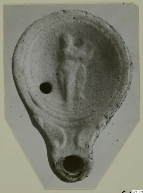 Terracotta oil lamp with a standing figure in relief on the disc, photographed on a grey card mount.