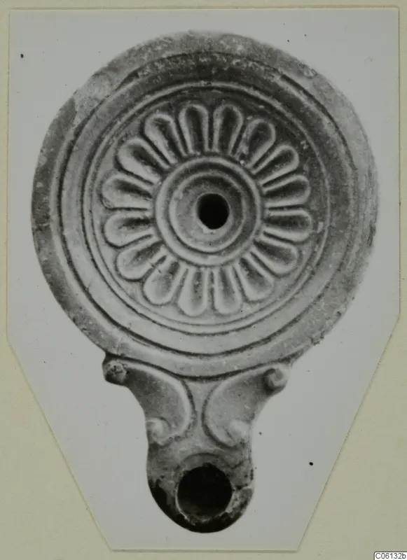 Terracotta oil lamp with a radiating petal pattern on its round disc and a handle at the base.