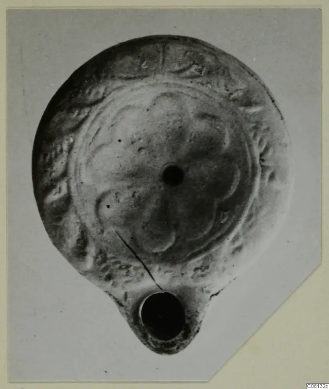 Terracotta oil lamp with a petal-like pattern on its disc, shown on a grey card mount.