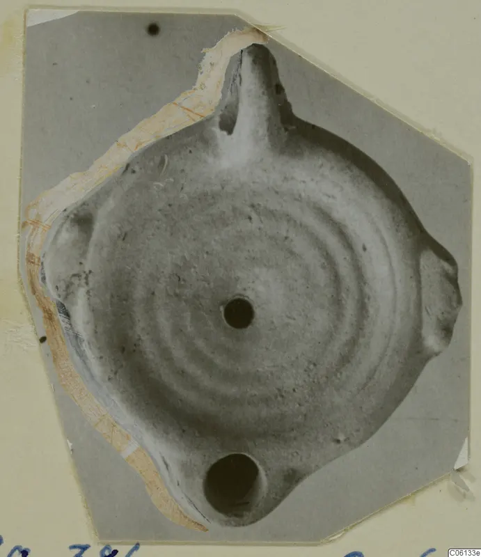 Terracotta lamp seen from above, with a nozzle at the top, a central filling hole and a handle, pasted on a grey card