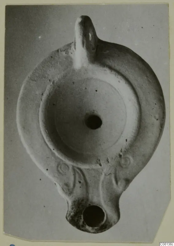 Terracotta oil lamp with a handle, seen from above, with a central filling hole and a nozzle at the bottom.