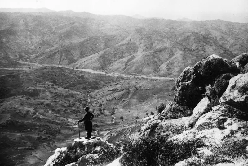 Wide view westward from a rocky outcrop over hills and a valley, with one standing man on the rocks in the foreground.