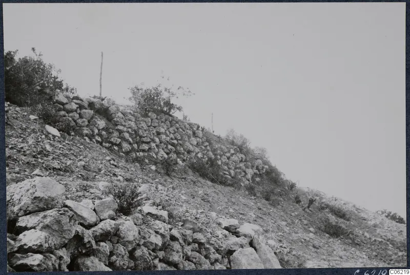 Rough stone wall running up a bare, stony hillside at Vouni, with scrub and a thin pole on the ridge.