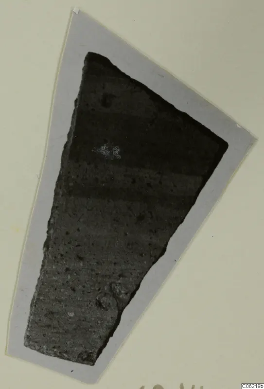 Wedge-shaped pottery sherd, dark grey with horizontal bands, shown on a grey card mount.
