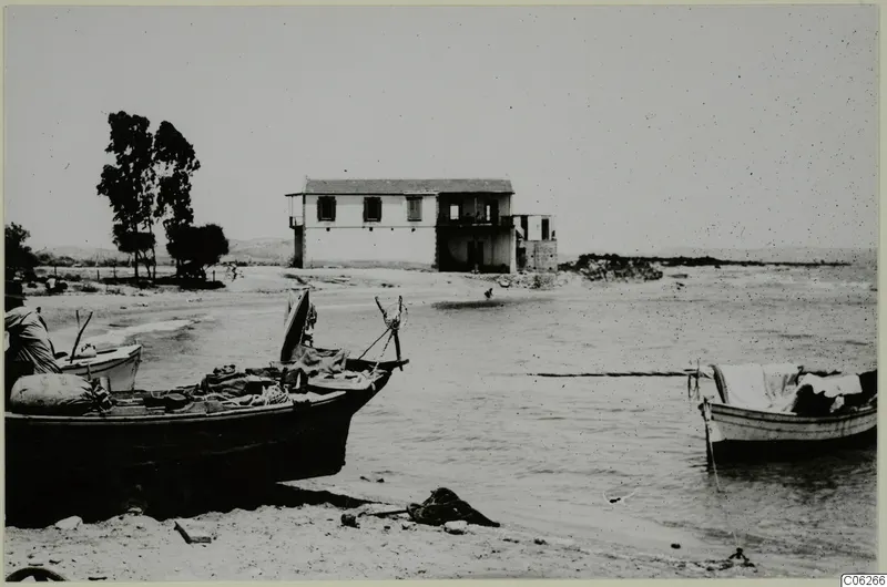 Boats drawn up on a sandy beach, with a two-storey whitewashed building and trees behind them