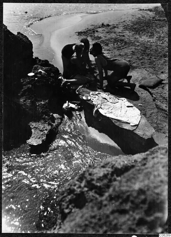 Three people crouched on sea-washed rocks at a Kyrenia beach, bending together over a small object.