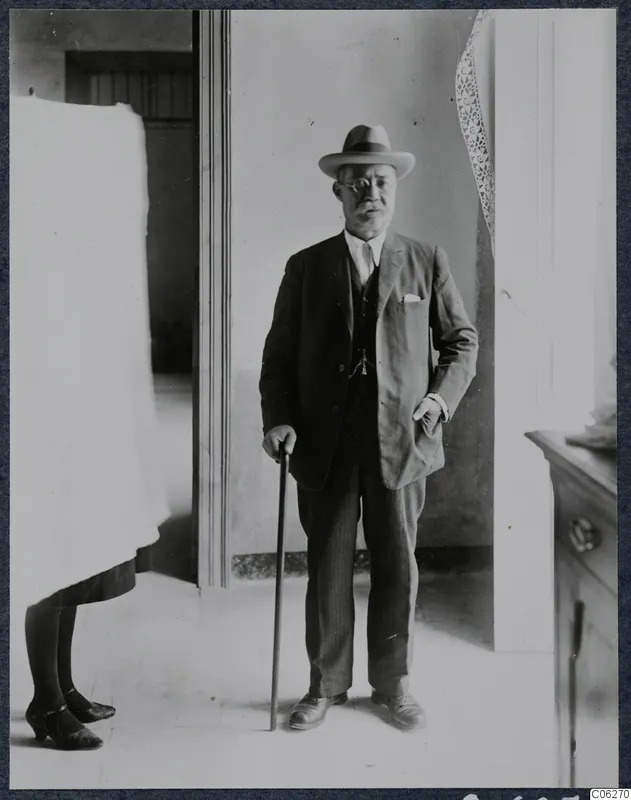Man in suit, hat and glasses holding a walking stick in a doorway, with part of another person visible at the left edge